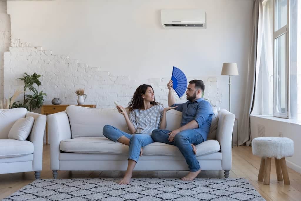 Couple sitting on their couch fanning themselves with AC unit hanging on the wall | Emergency Heating and AC Repair Services