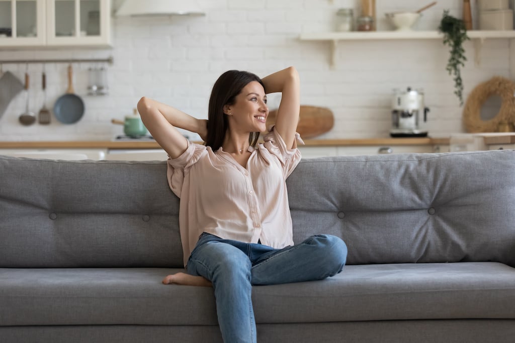 Woman sitting on a grey couch in her home smiling and looking to the side | Indoor Air Quality Services in Dallas, Frisco, and Plano, TX