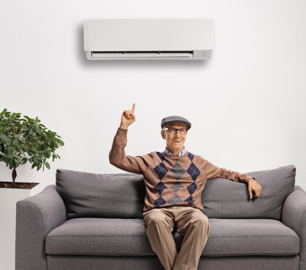 Elderly man seated on a sofa pointing up towards an air conditioning unit on the wall. AC Repair in Farmers Branch